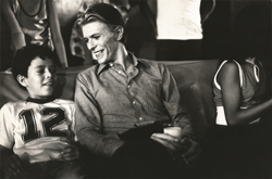 thumbnail link to original press photograph David Bowie on set 1975, on sofa next to boy, with glass of milk.